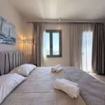 Monte Vista Lemnos Apartments suite with a king-size bed, soft grey curtains, sea-view windows and neatly rolled towels in a calm, airy interior.