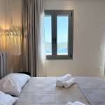 A cozy Monte Vista Lemnos Apartments bedroom with neutral bedding, rolled towels, and a window overlooking the blue sea and green hills in Lemnos.