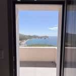 Open doorway from Monte Vista Lemnos Apartments frames a balcony and sweeping bay view with blue sea, hills, and a clear sky over the coast.
