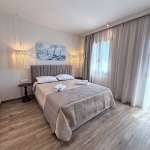Monte Vista bedroom with a king-size bed, soft beige decor, sea-themed wall art, pendant lights, and floor-to-ceiling curtains beside a bright window.