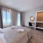 Bright modern bedroom at Monte Vista Lemnos Apartments with a double bed, sheer curtains, work desk, round mirror, wood flooring, and minimalist decor.