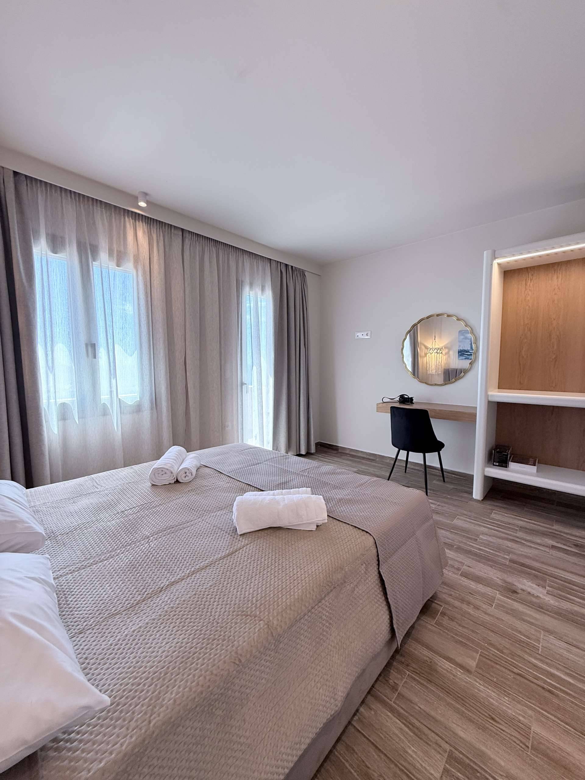 Bright modern bedroom at Monte Vista Lemnos Apartments with a double bed, sheer curtains, work desk, round mirror, wood flooring, and minimalist decor.