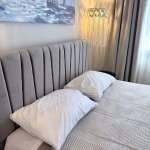 Monte Vista Lemnos Apartments bedroom with a grey upholstered headboard, white pillows, soft curtains and a sailboat painting by the bed.