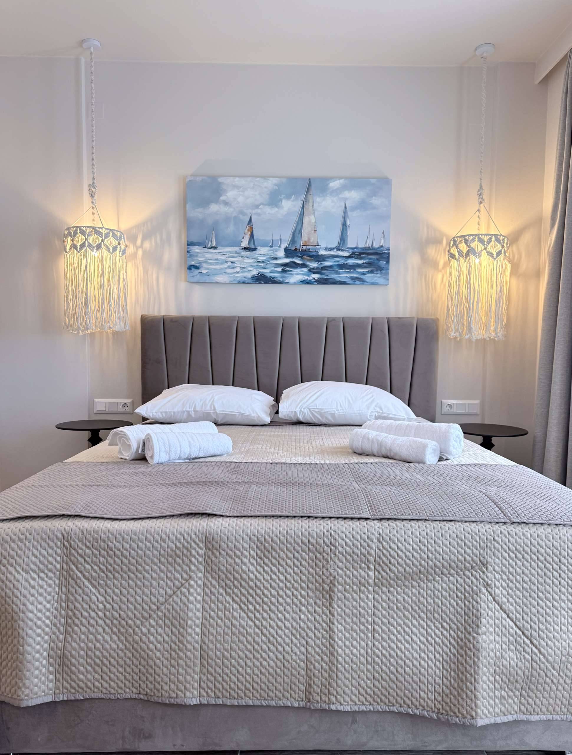 A softly lit Monte Vista Lemnos Apartments bedroom with a gray upholstered bed, sailboat painting, bedside tables, and woven pendant lights.