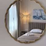 A gold-framed round mirror reflects a calm Monte Vista Lemnos bedroom with a neatly made bed, sheer curtains, warm lamp light and sailboat art.