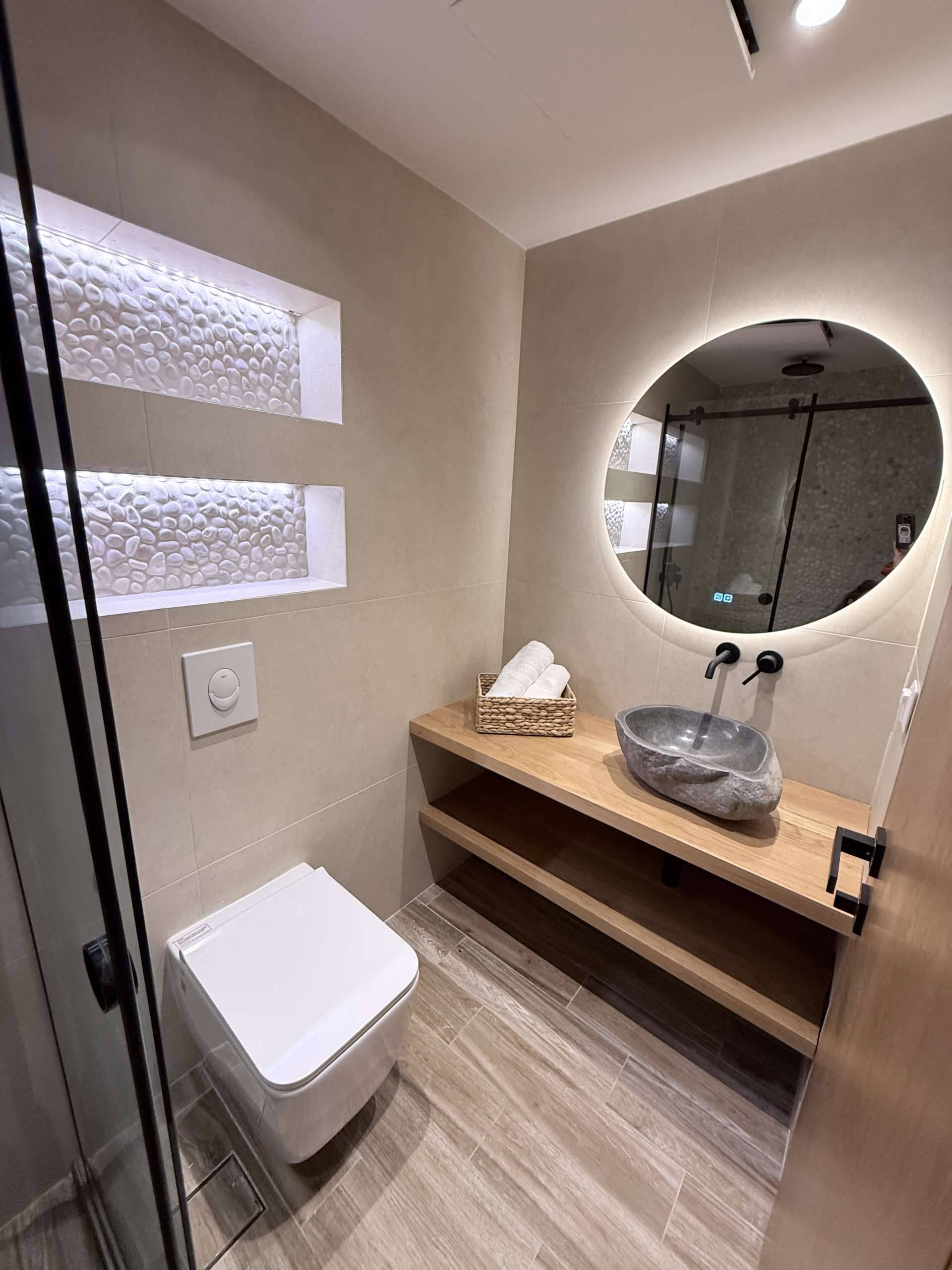 Modern bathroom with a stone vessel sink, round backlit mirror, wooden vanity, glass shower, and wall niches in Monte Vista Lemnos Apartments.
