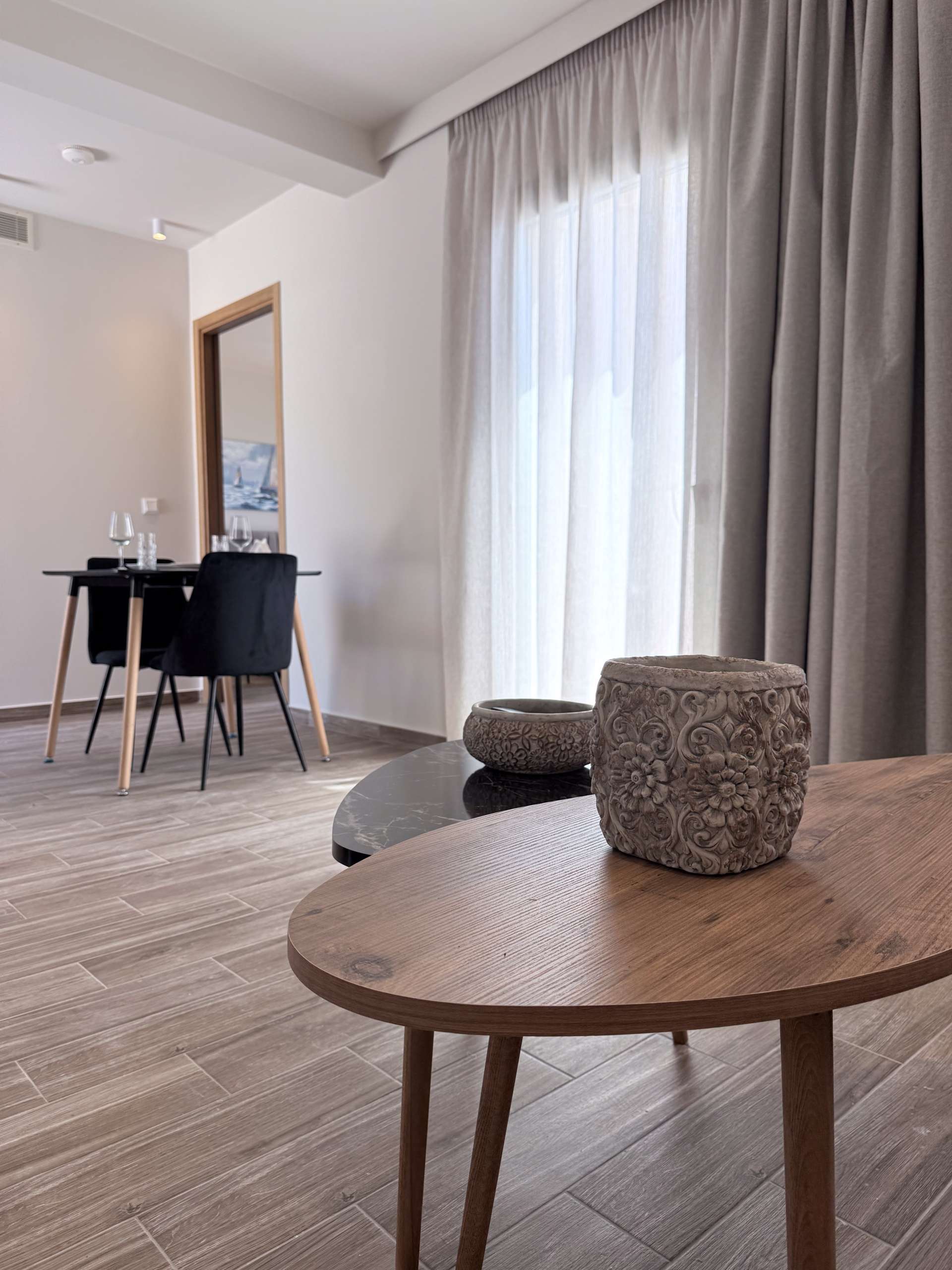 Bright Monte Vista Lemnos Apartments interior with a wooden coffee table, black dining chairs, soft gray curtains, and natural light over light wood flooring.
